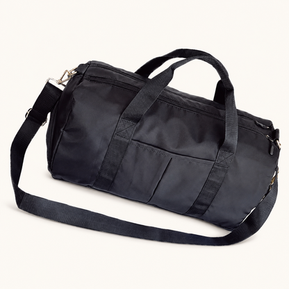 Black Split Duffel Bag with carry handles, shoulder strap and front slip pockets on a warm off-white studio background.