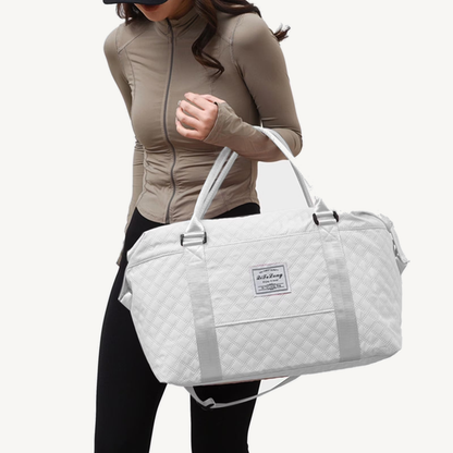 White quilted weekender duffel with long carry handles and shoulder strap, carried by a woman against a warm off-white background.